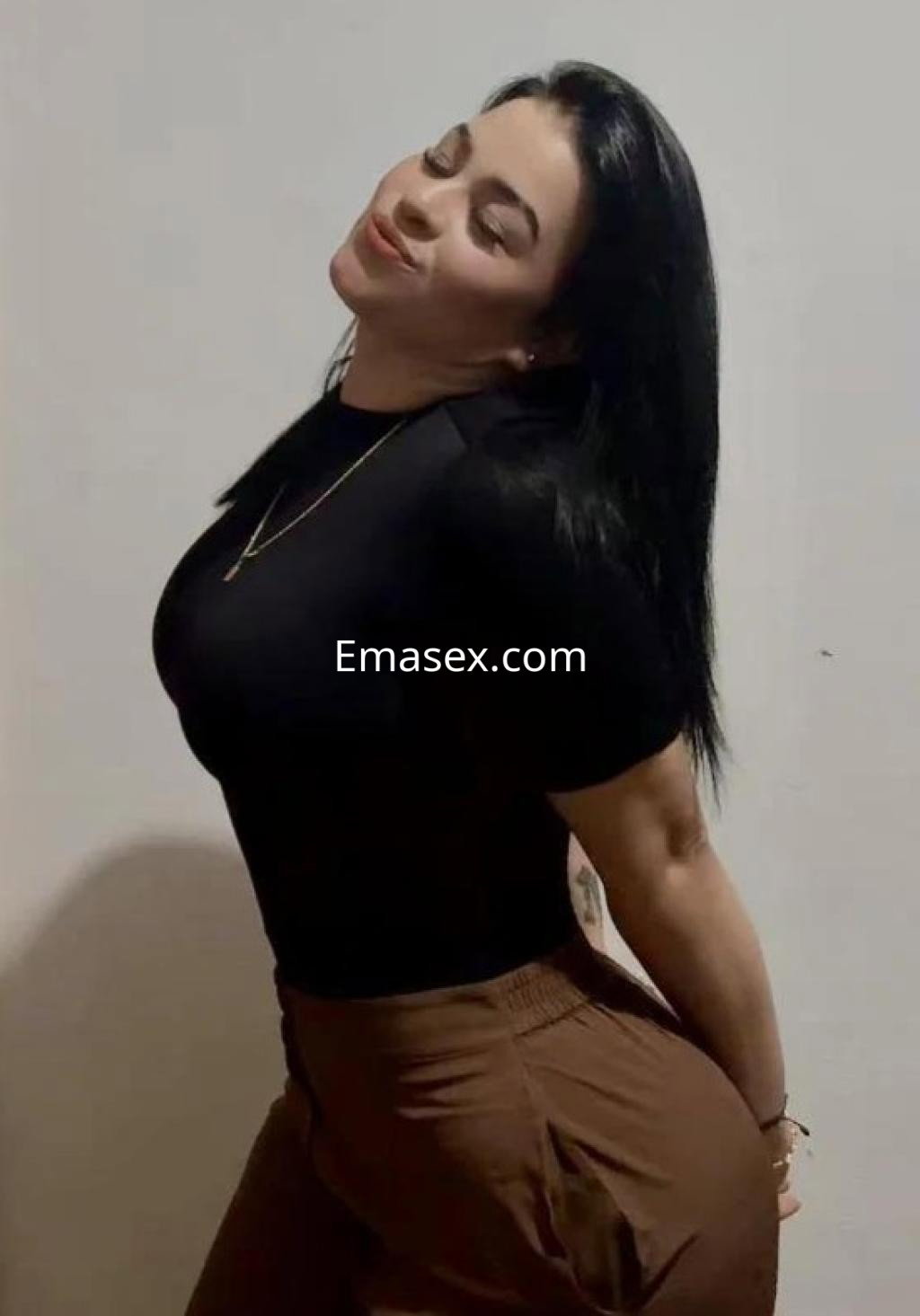 Puta en Girona, con cabello negro y lacio, viste una camiseta negra y pantalones marrones, sonriendo y posando.