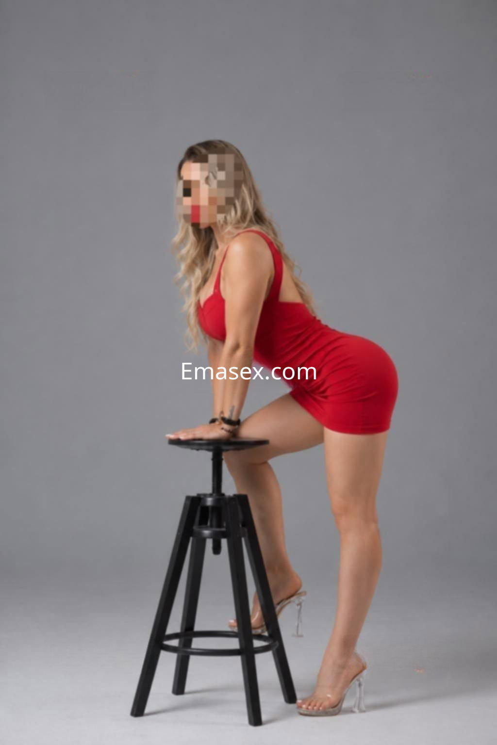 Escort en Valencia posando de perfil sobre un taburete, vistiendo un vestido rojo y tacones transparentes.