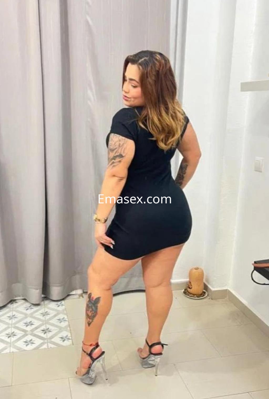 Escort en Toledo posando de espaldas con un vestido negro ajustado y tacones transparentes, mostrando un tatuaje en su pierna.