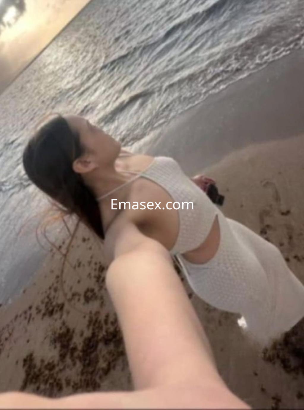 Puta en Valencia, de pie en la playa con un vestido blanco, mirando hacia el mar en un ambiente relajado.