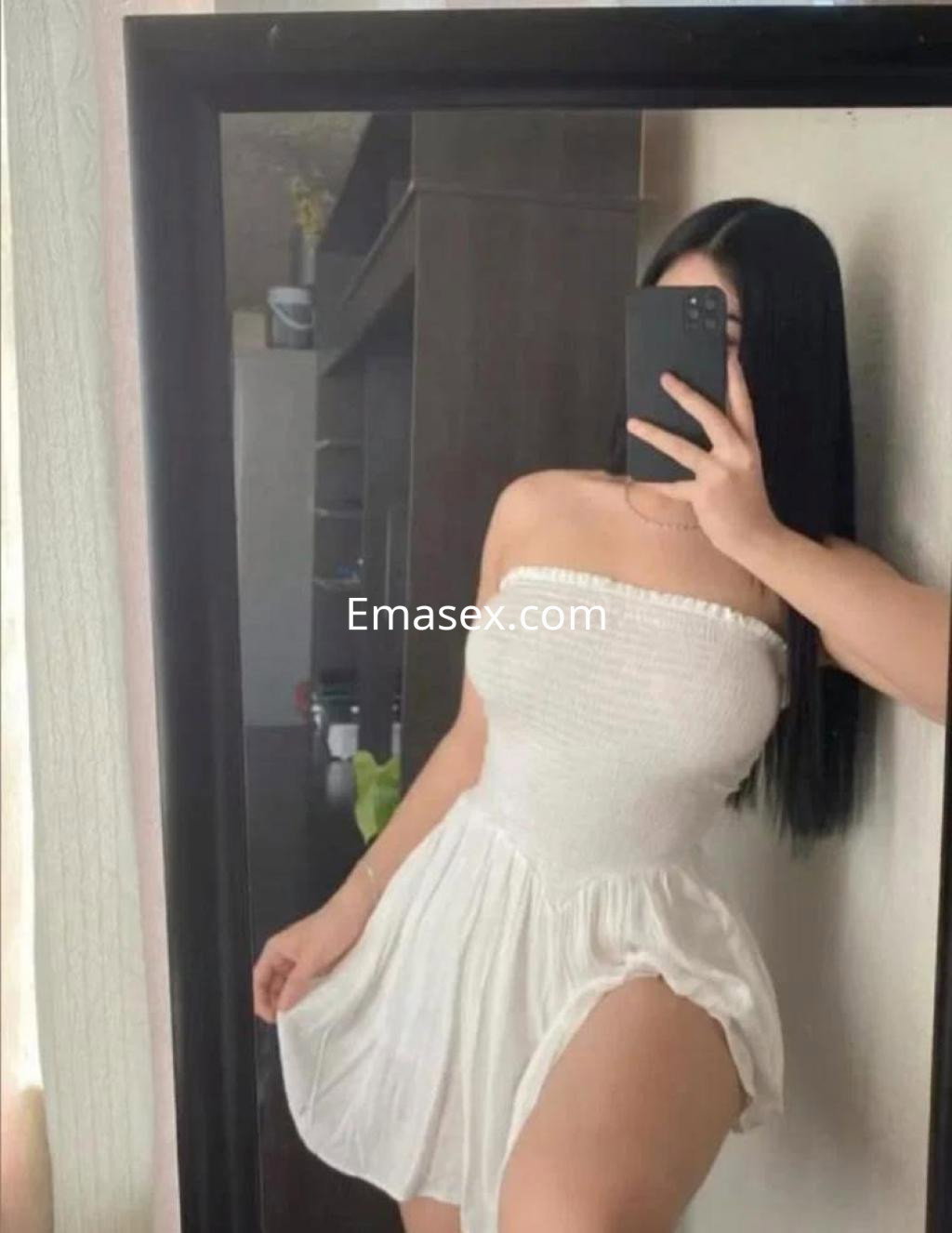 Escort en Palma de Mallorca, de pie frente a un espejo, con un vestido blanco sin mangas y cabello lacio y oscuro.