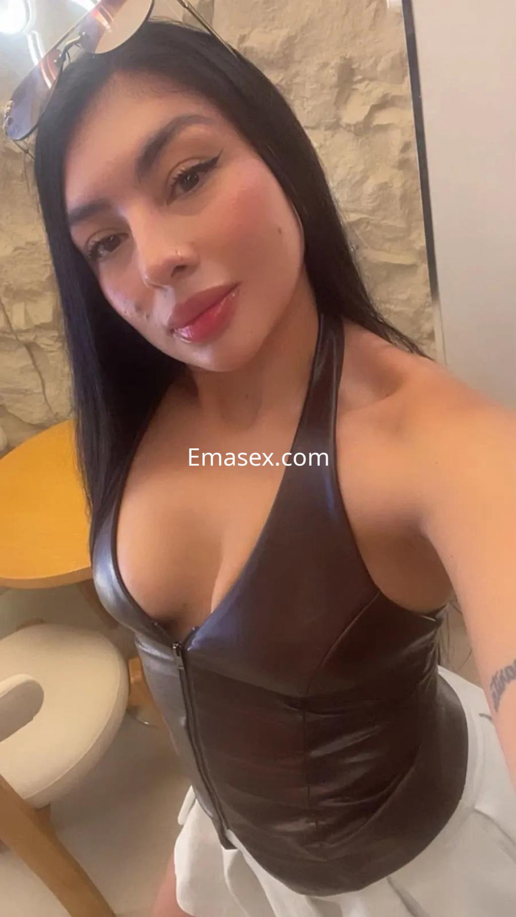 Escort en Valencia con blusa de cuero ajustada y falda blanca, posando con una sonrisa en un entorno acogedor.