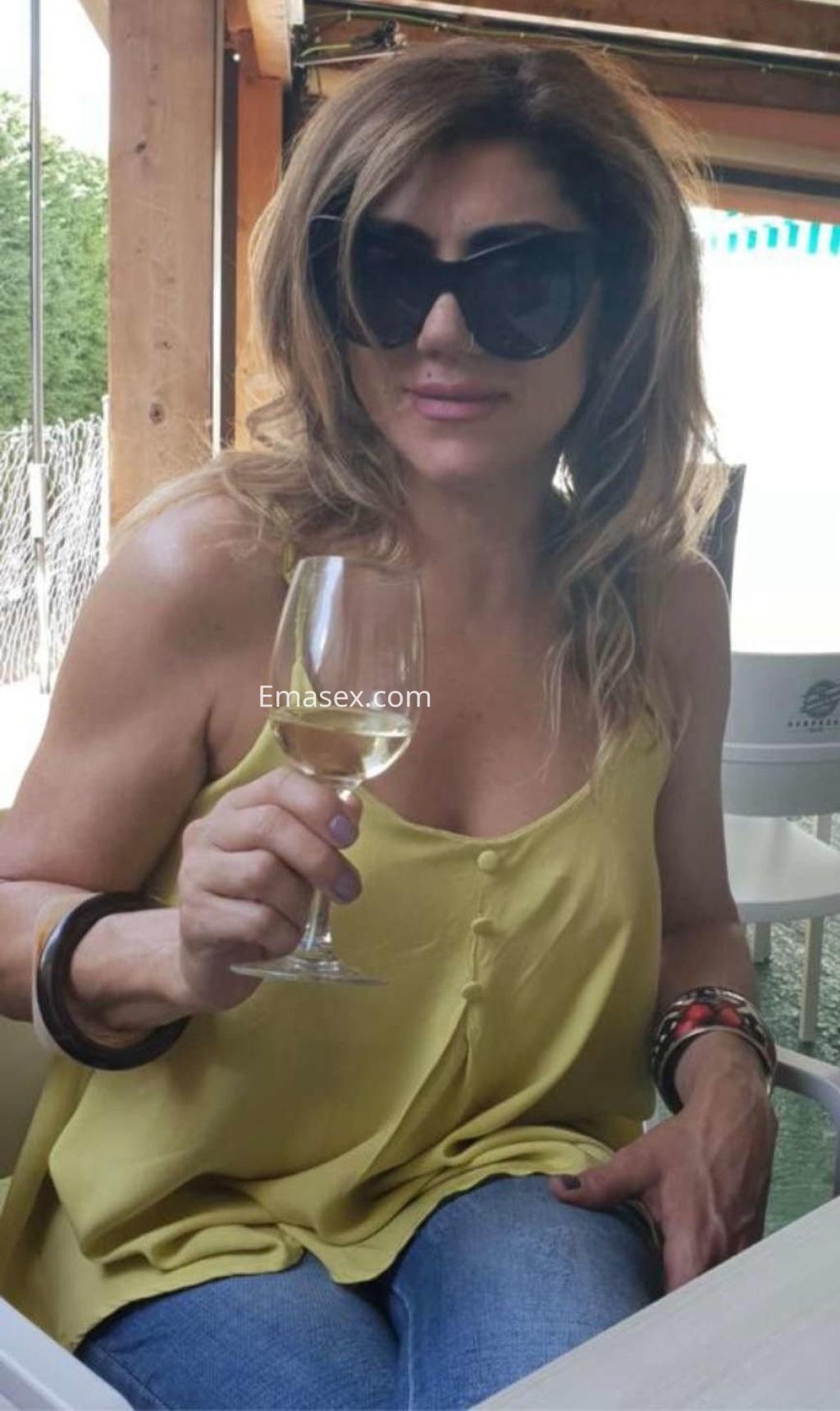 Escort en Toledo con cabello ondulado y castaño claro, luciendo una blusa amarilla y sosteniendo una copa de vino.