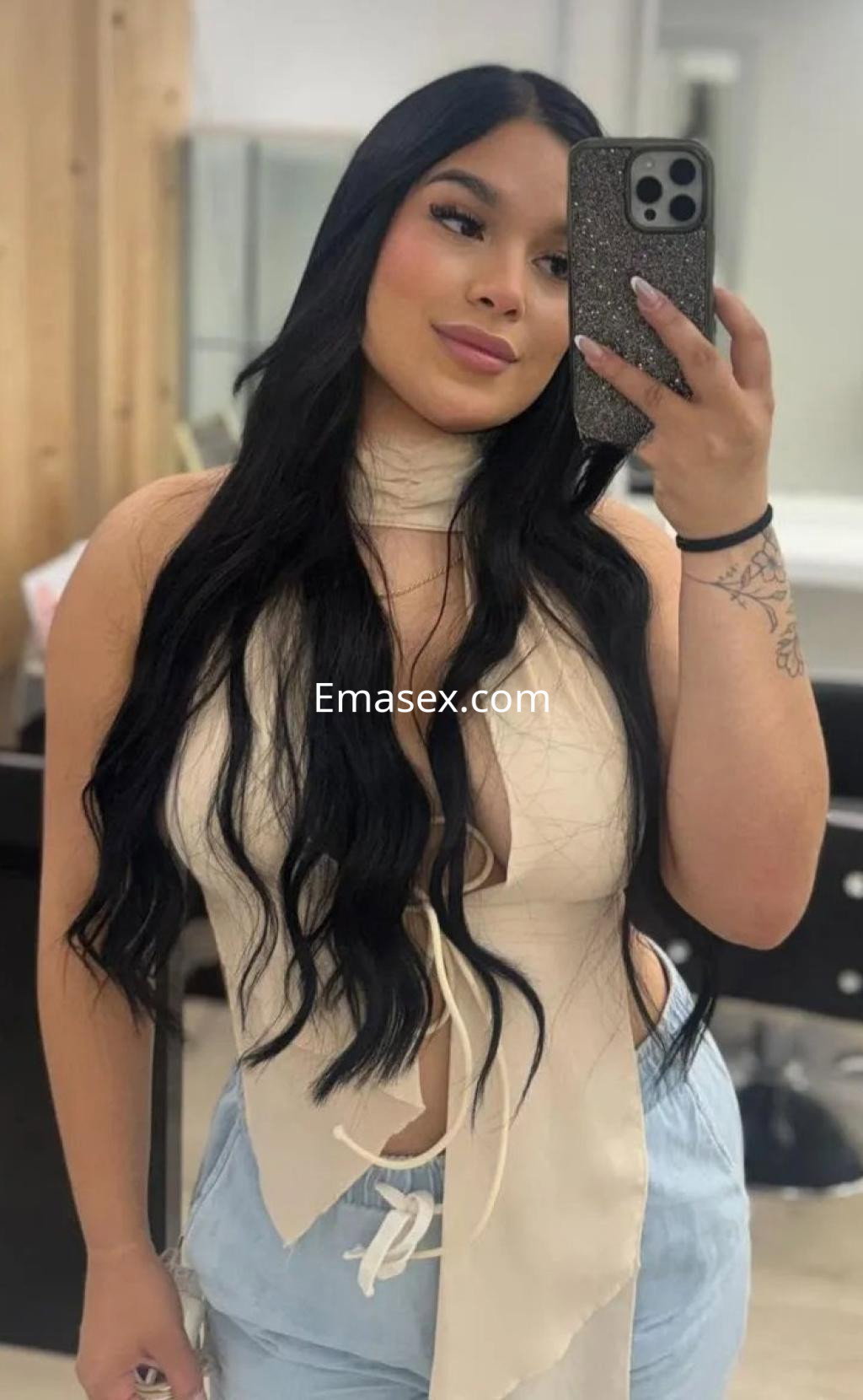 Puta en Valencia con cabello largo y ondulado, viste una blusa clara con escote y pantalones claros, posando en un espejo.