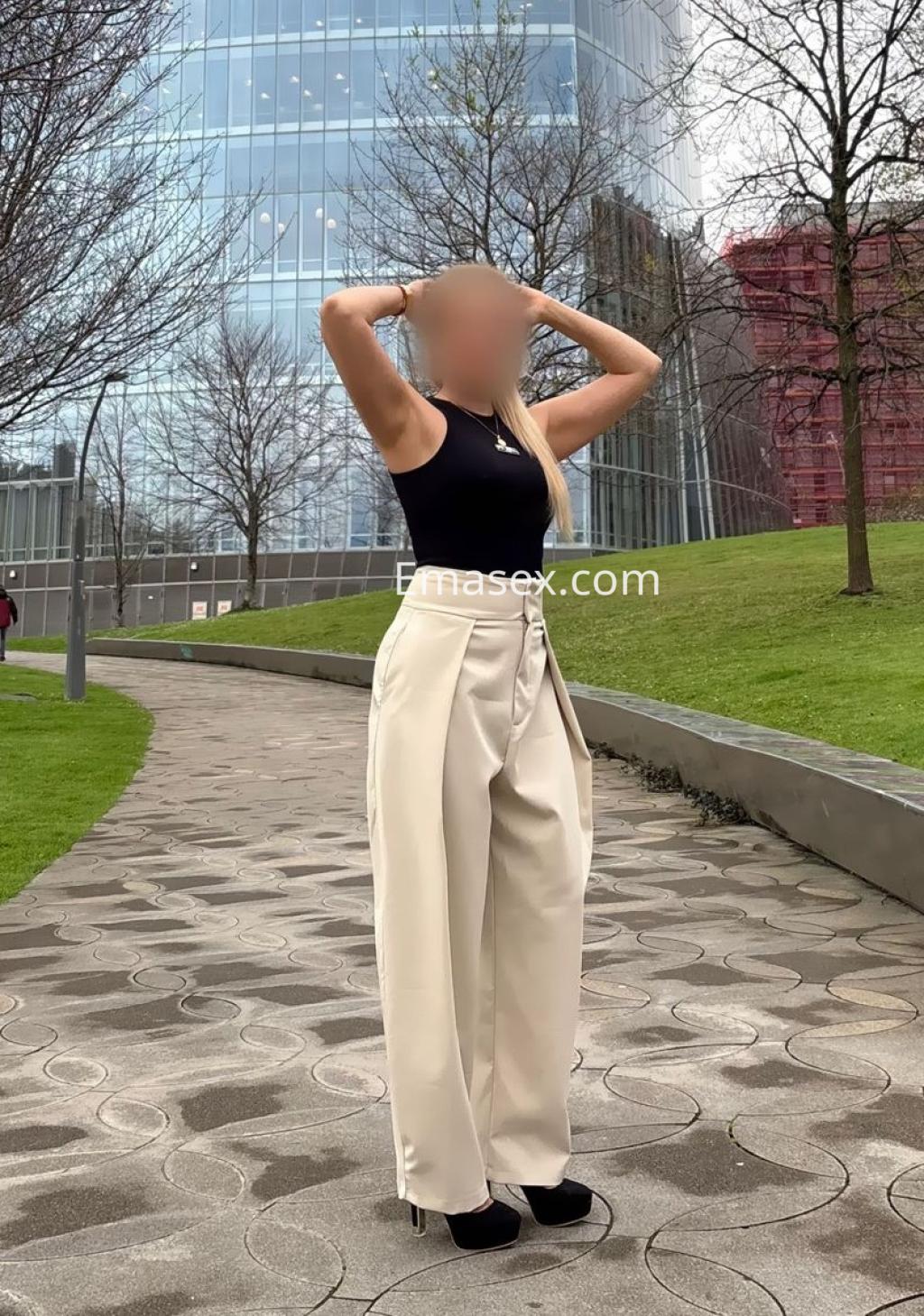 Puta con cabello rubio, viste una blusa negra y pantalones claros, posando en un parque urbano de Bilbao.