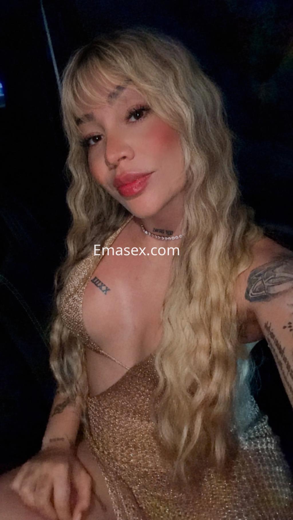 Escort en Logroño con cabello rubio y ondulado, vistiendo un vestido brillante y posando en un ambiente oscuro.
