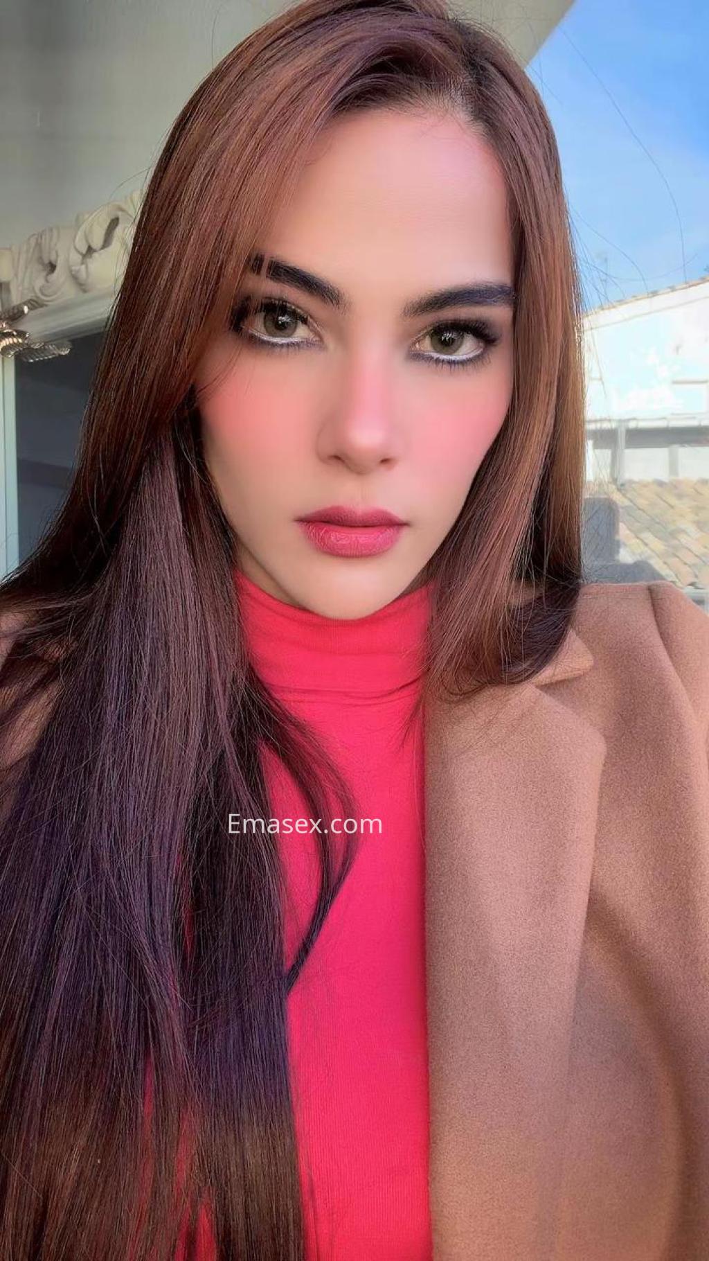 La escort en Lleida aparece en un primer plano, con cabello largo y castaño, vistiendo un suéter rojo y abrigo beige.