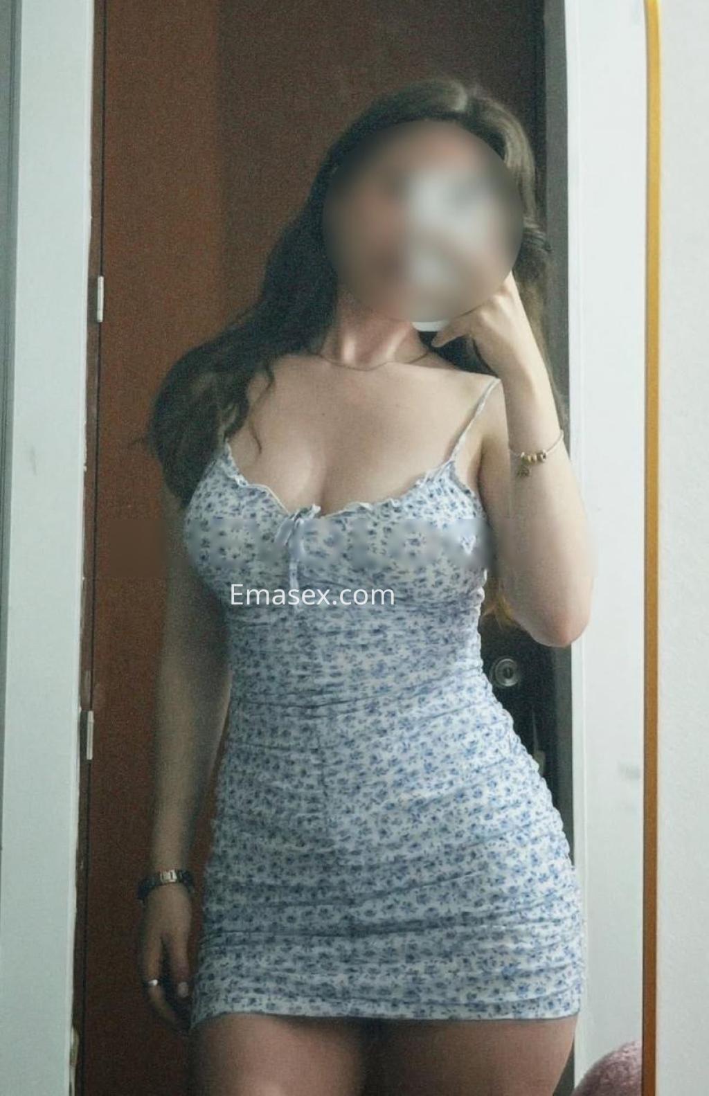 Puta en Lleida posando frente al espejo con un vestido ajustado de flores y un peinado ondulado, sonriendo.
