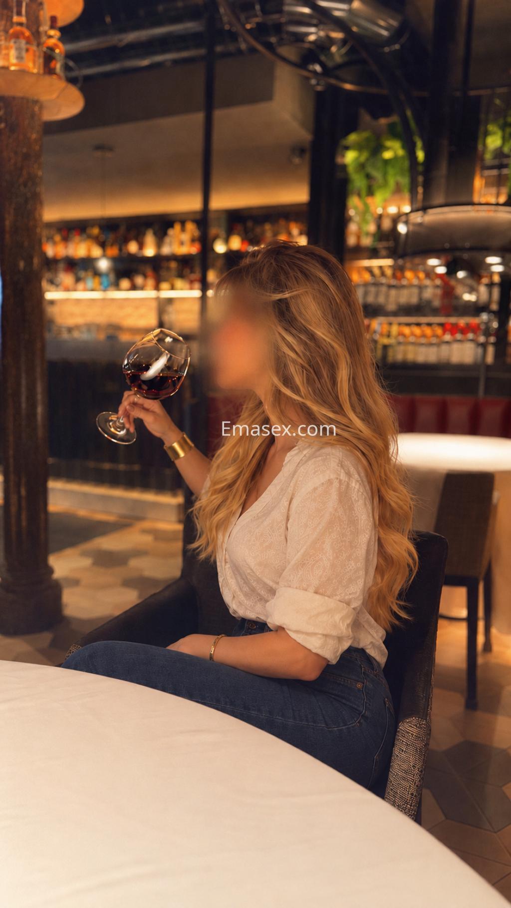 Escort en Bilbao, sentada en una mesa con una blusa clara y jeans, sosteniendo una copa de vino en un ambiente elegante.