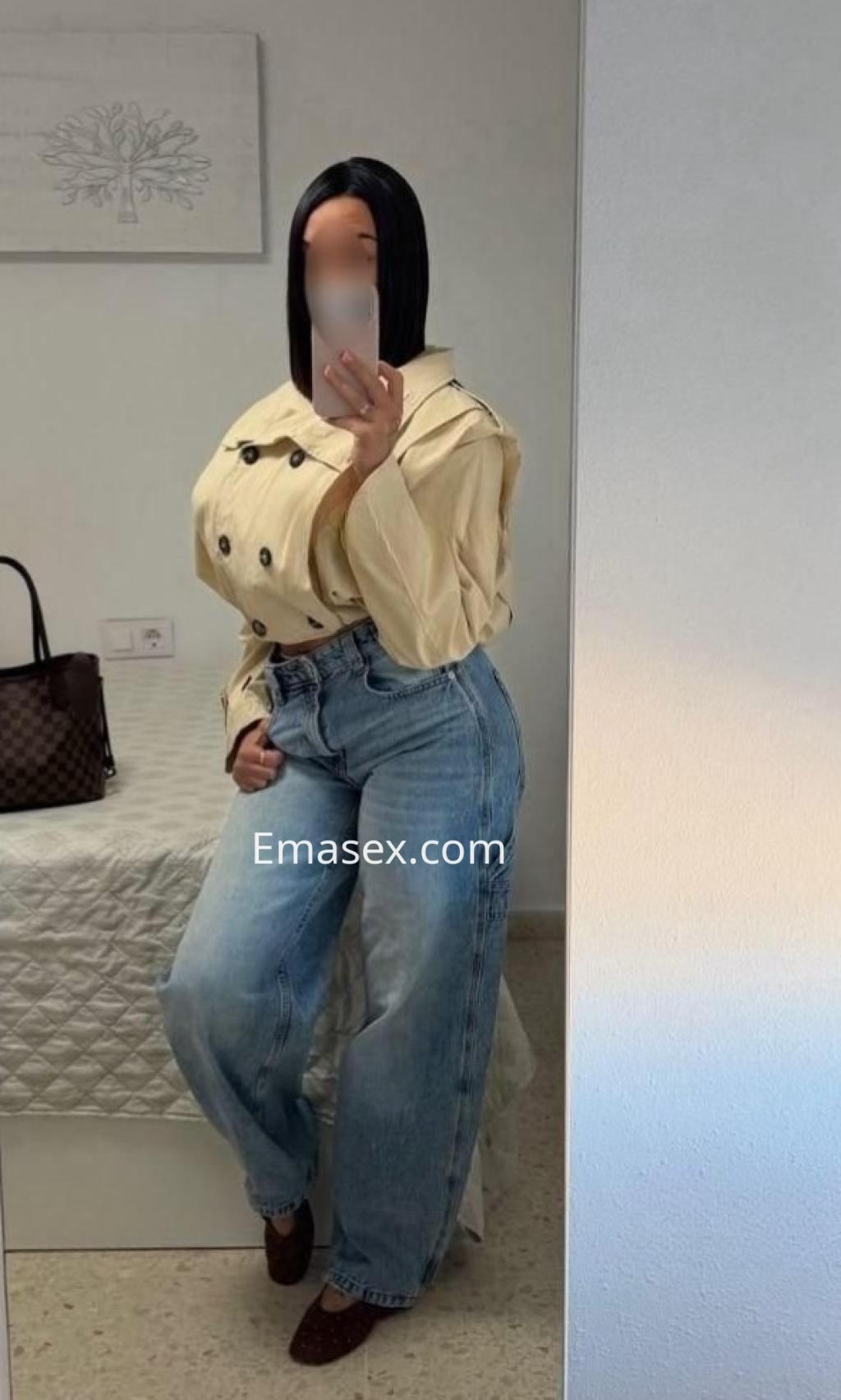 Puta en Cádiz posando frente a un espejo, con cabello lacio y oscuro, blusa clara y jeans anchos.