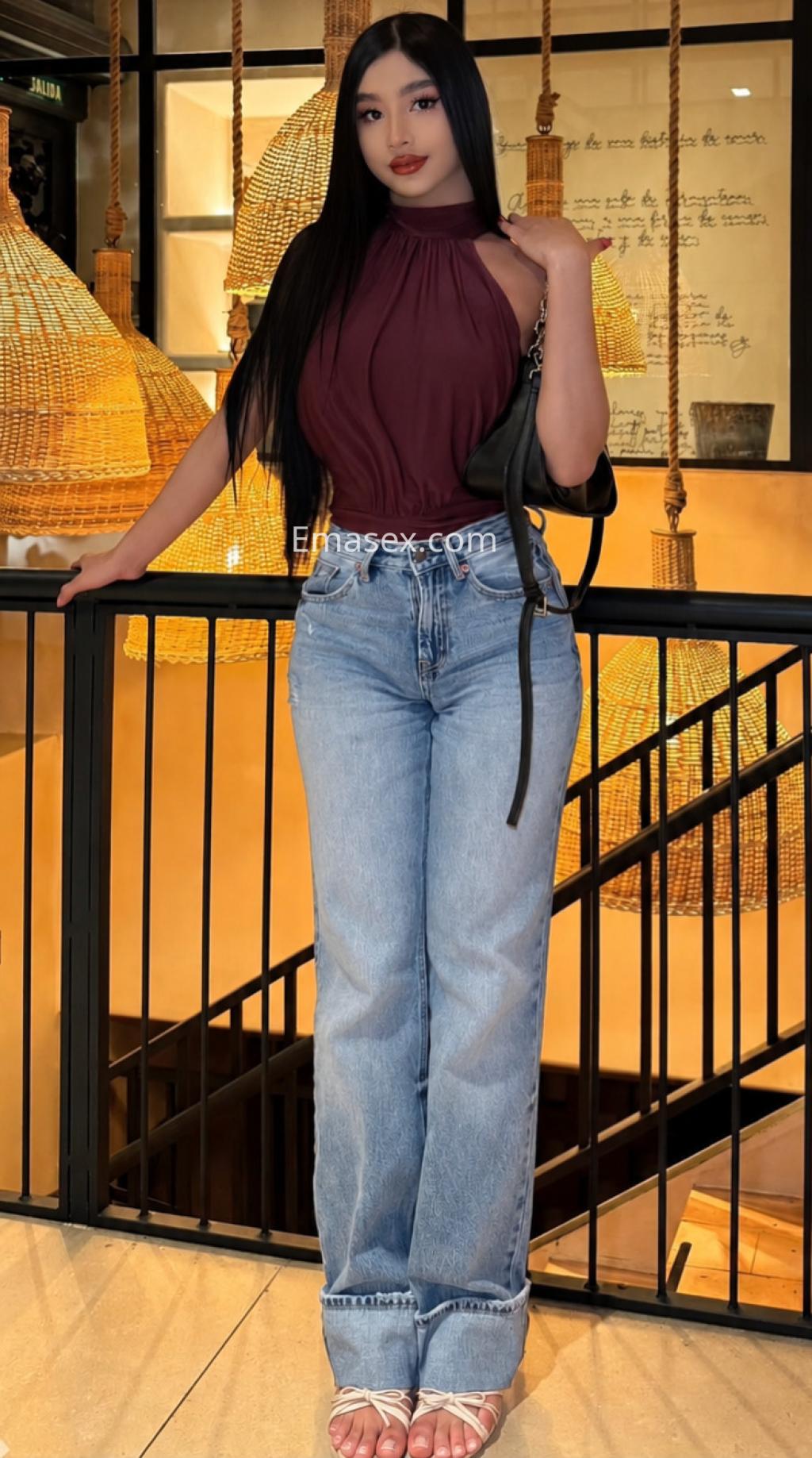 Escort trans en Logroño, con cabello largo y lacio, vistiendo una blusa burdeos y jeans, posando en un ambiente moderno.