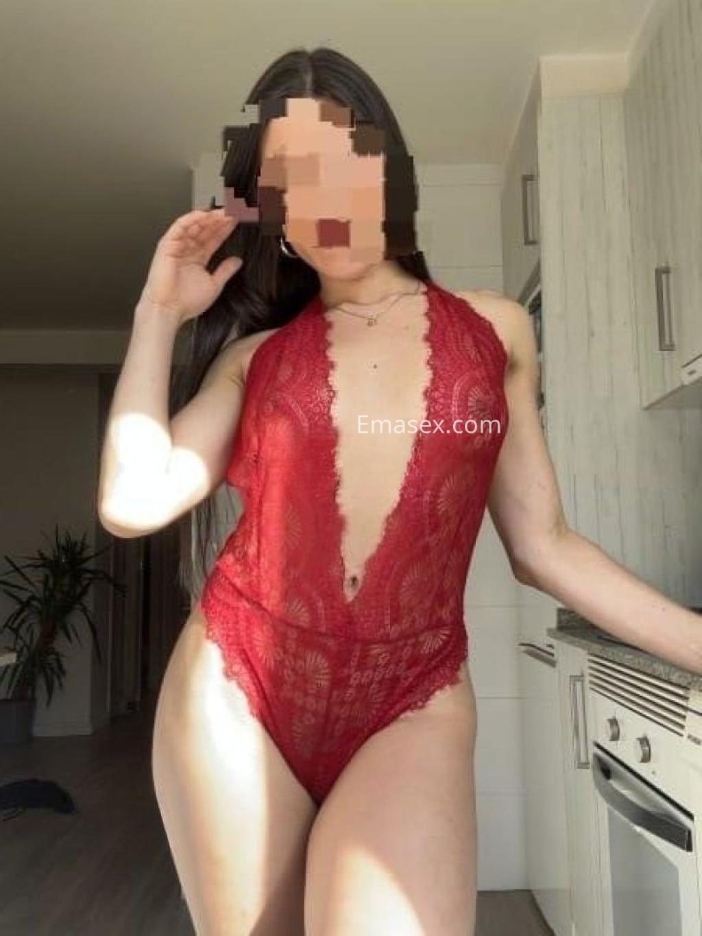 Escort en Pontevedra viste un body de encaje rojo con escote pronunciado, posando en una cocina luminosa.