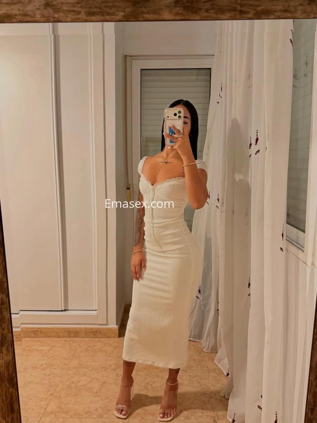 Escort en Alicante con vestido blanco ajustado, tacones y cabello oscuro, posando frente a un espejo.