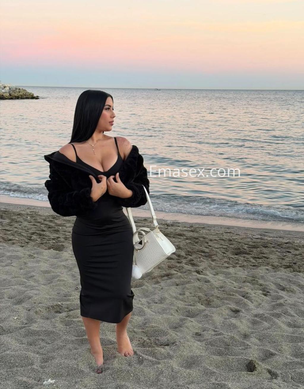 Puta de cabello oscuro, con vestido negro ajustado y chaqueta de piel, posando en la playa de Málaga al atardecer.