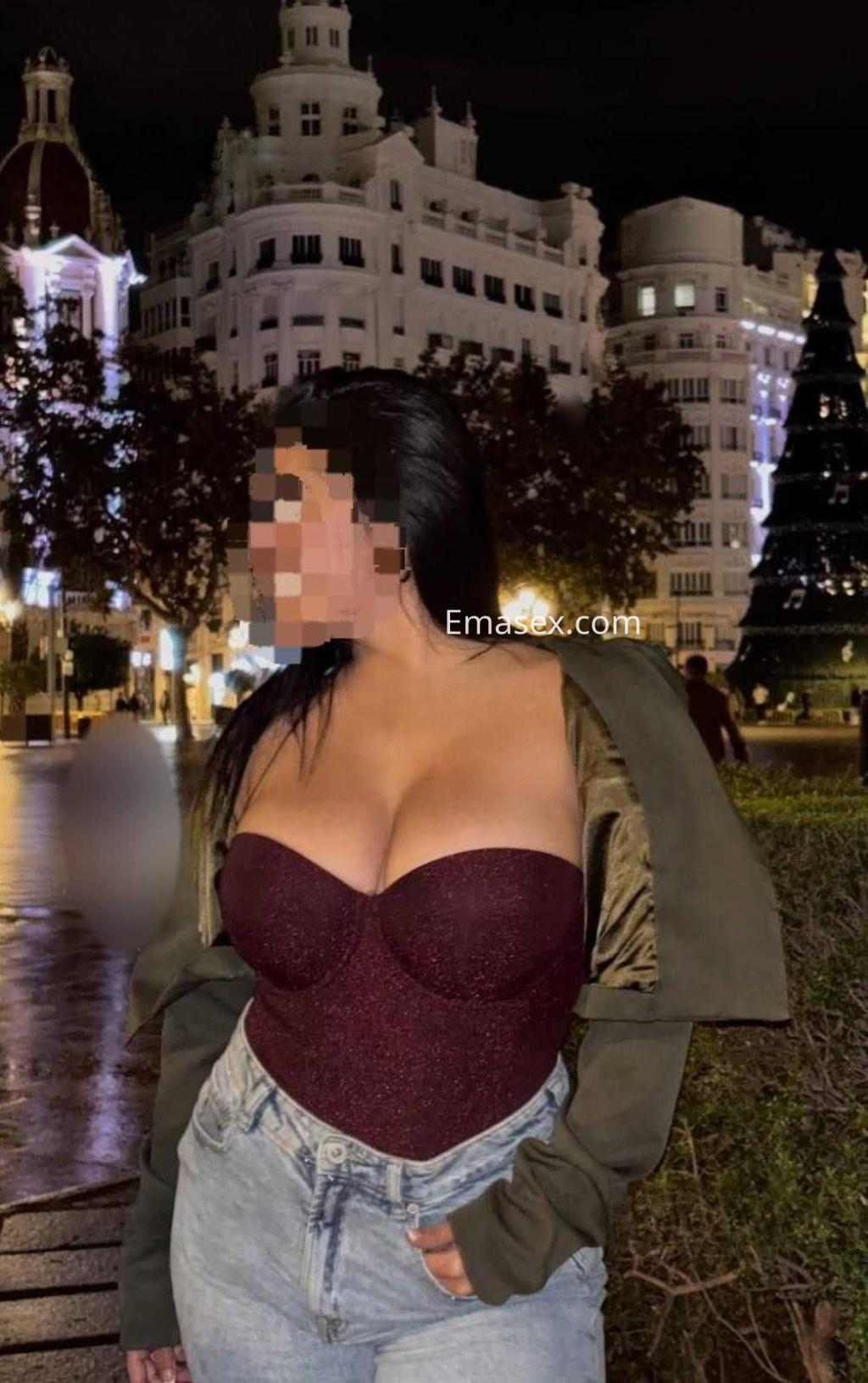 Puta de complexión curvilínea viste un body burdeos y chaqueta, posando en la iluminada Alicante nocturna.
