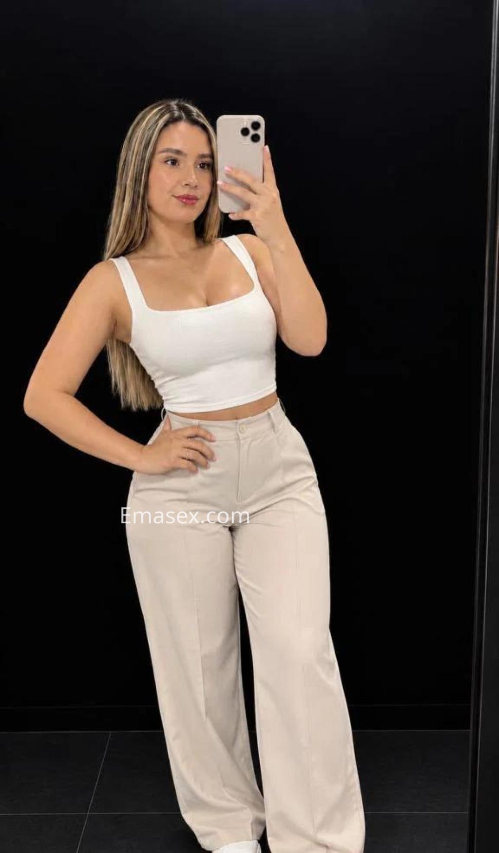 Escort en Madrid con blusa blanca ajustada y pantalones claros, posando frente a un espejo.