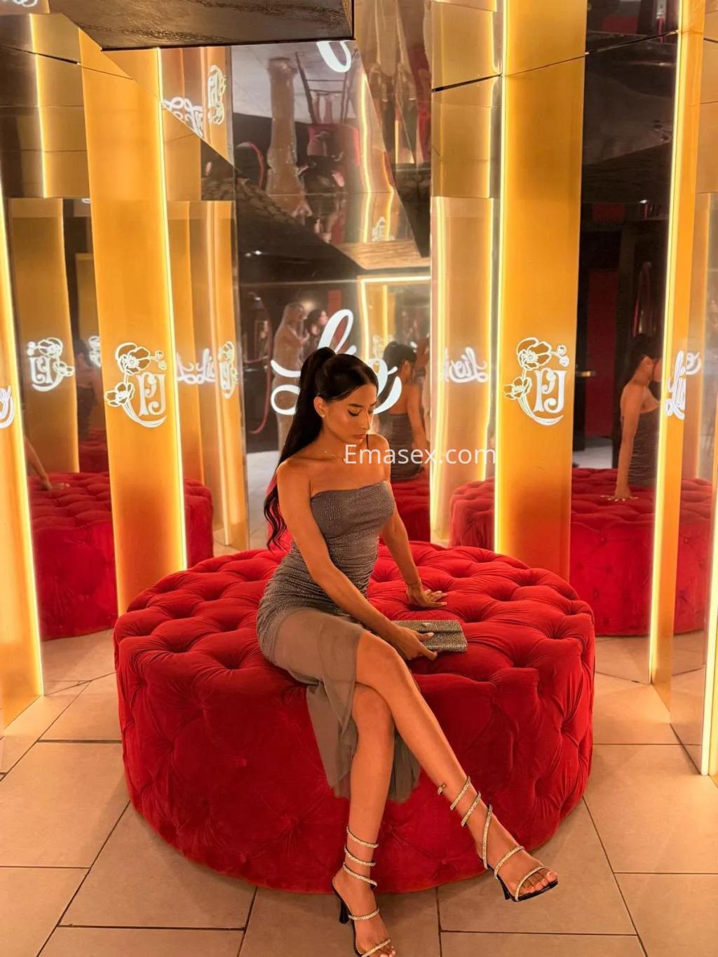Puta posando en una silla roja en Coruña, vistiendo un vestido ajustado y sandalias de tiras, rodeada de espejos.