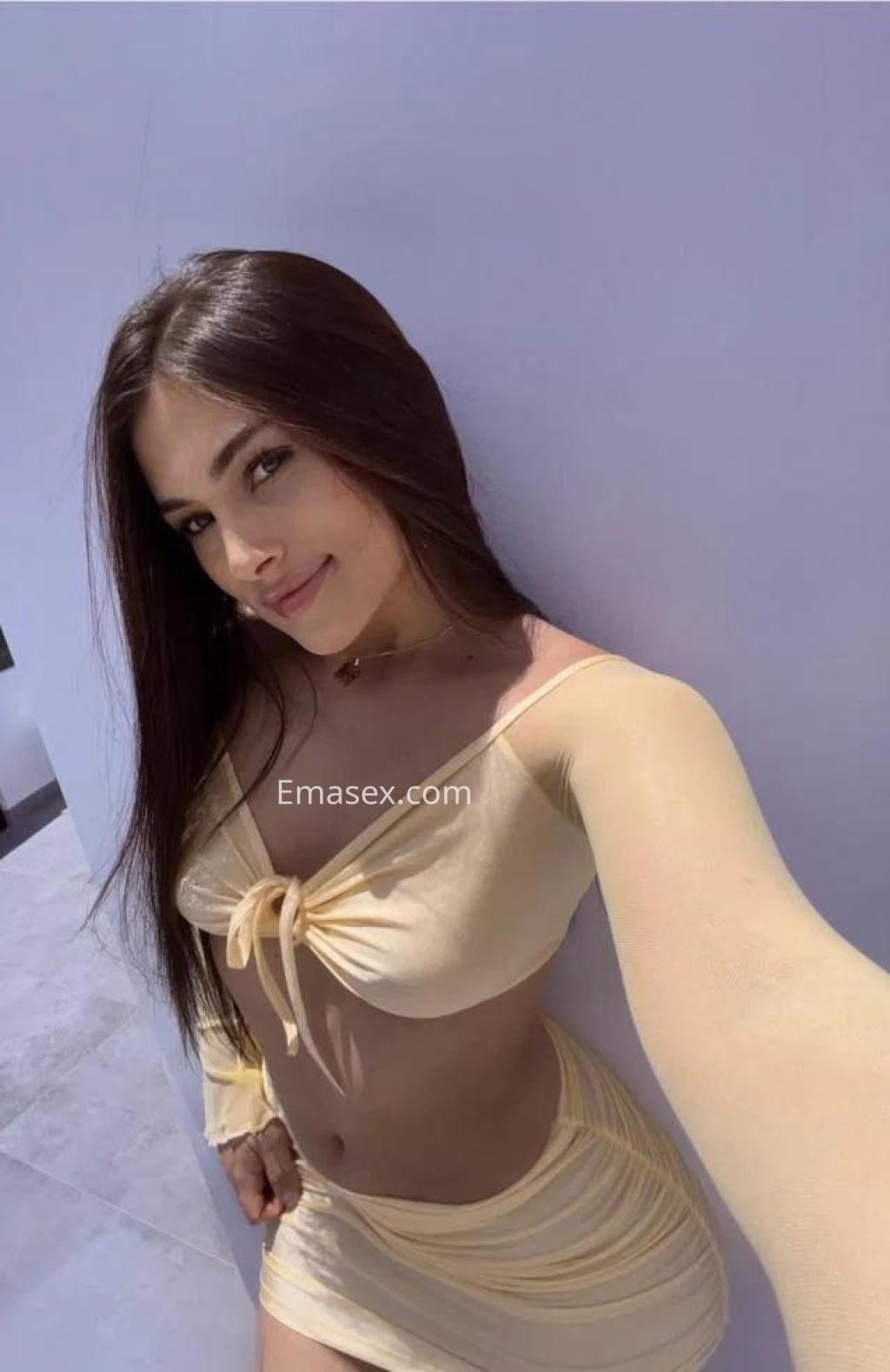Puta de Lleida con blusa amarilla ajustada y falda corta, sonriendo frente a una pared clara.