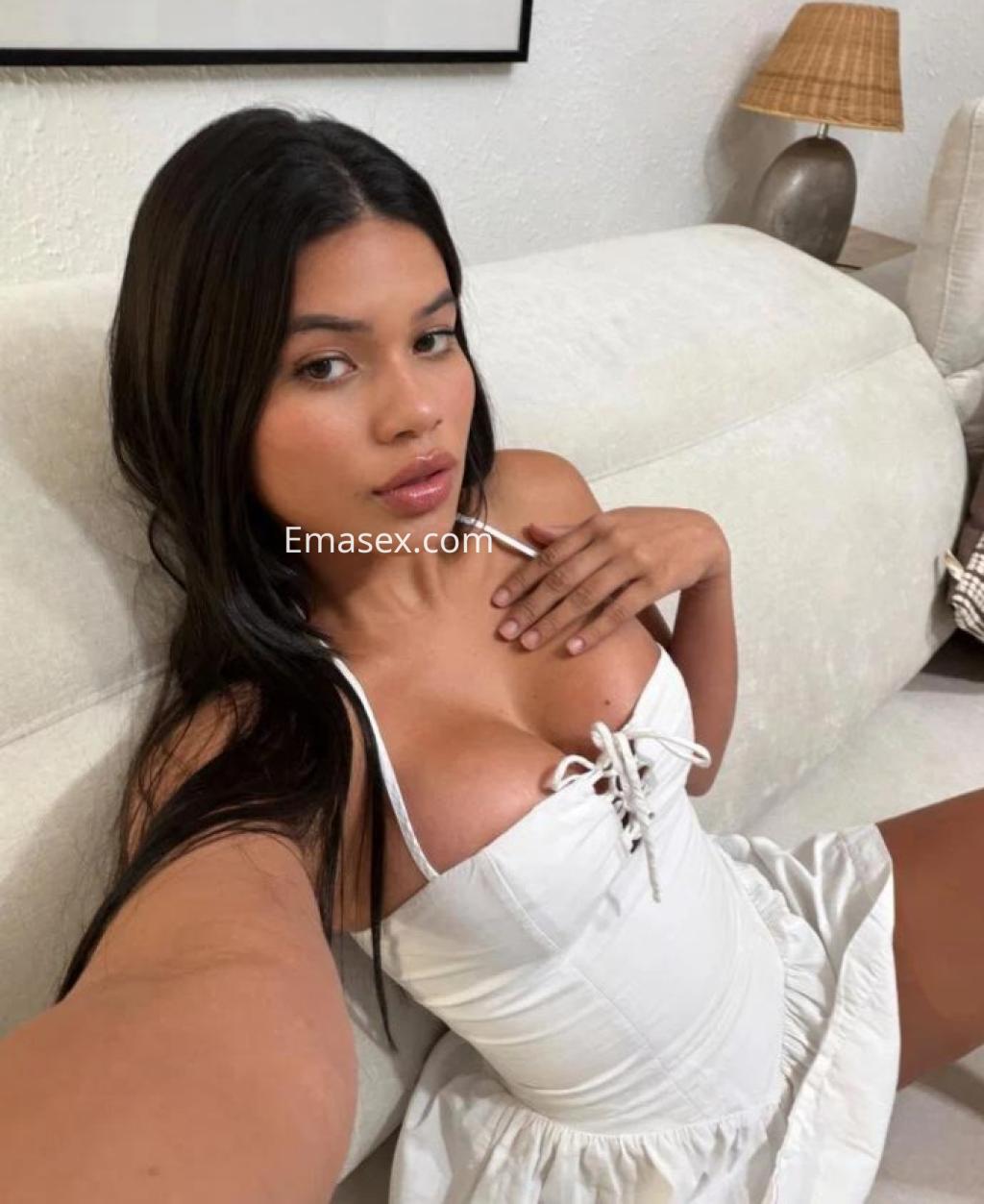 Escort en Málaga con cabello largo y oscuro, vistiendo un vestido blanco, posando sentada en un sofá claro.