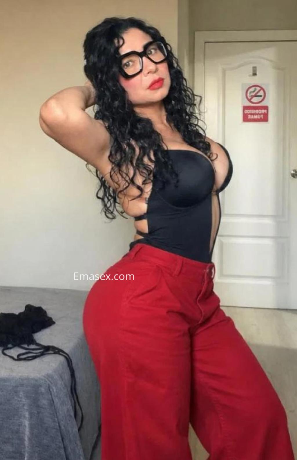 Escort con cabello rizado y oscuro, vistiendo un top negro y pantalones rojos, posando en un ambiente interior en Madrid.