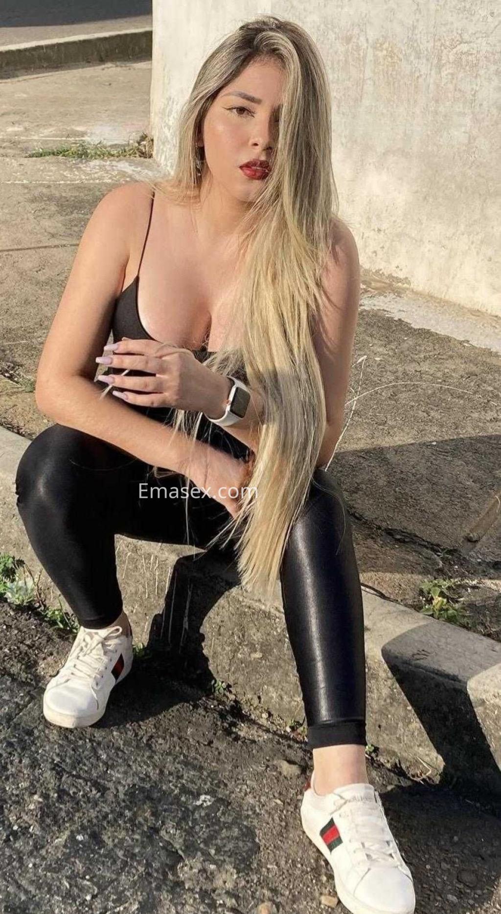 Escort en Madrid, con cabello largo y rubio, viste un top negro y leggings brillantes, sentada en una acera.