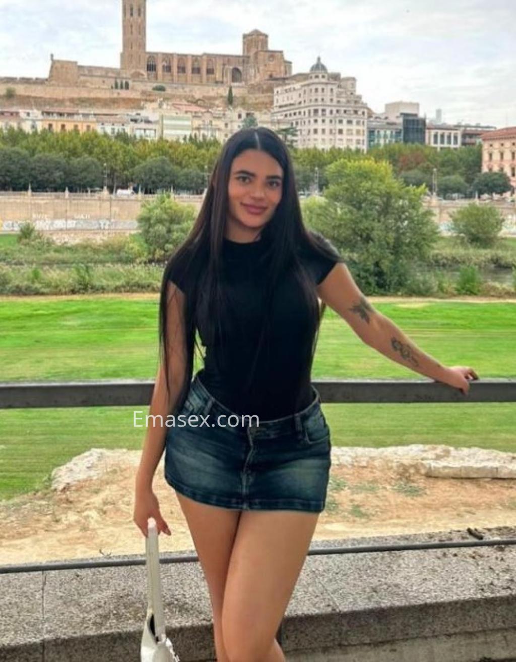 Puta en Lleida, posando con una camiseta negra y una mini falda de mezclilla, con un paisaje urbano de fondo.