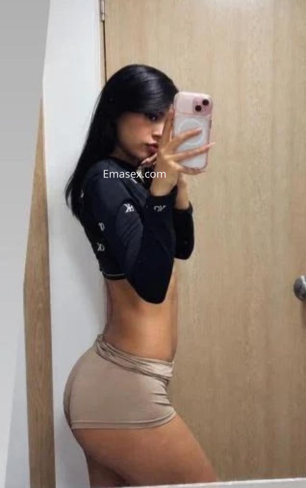 La puta en Málaga se fotografía de perfil en el espejo, vistiendo un top negro y una falda corta beige.