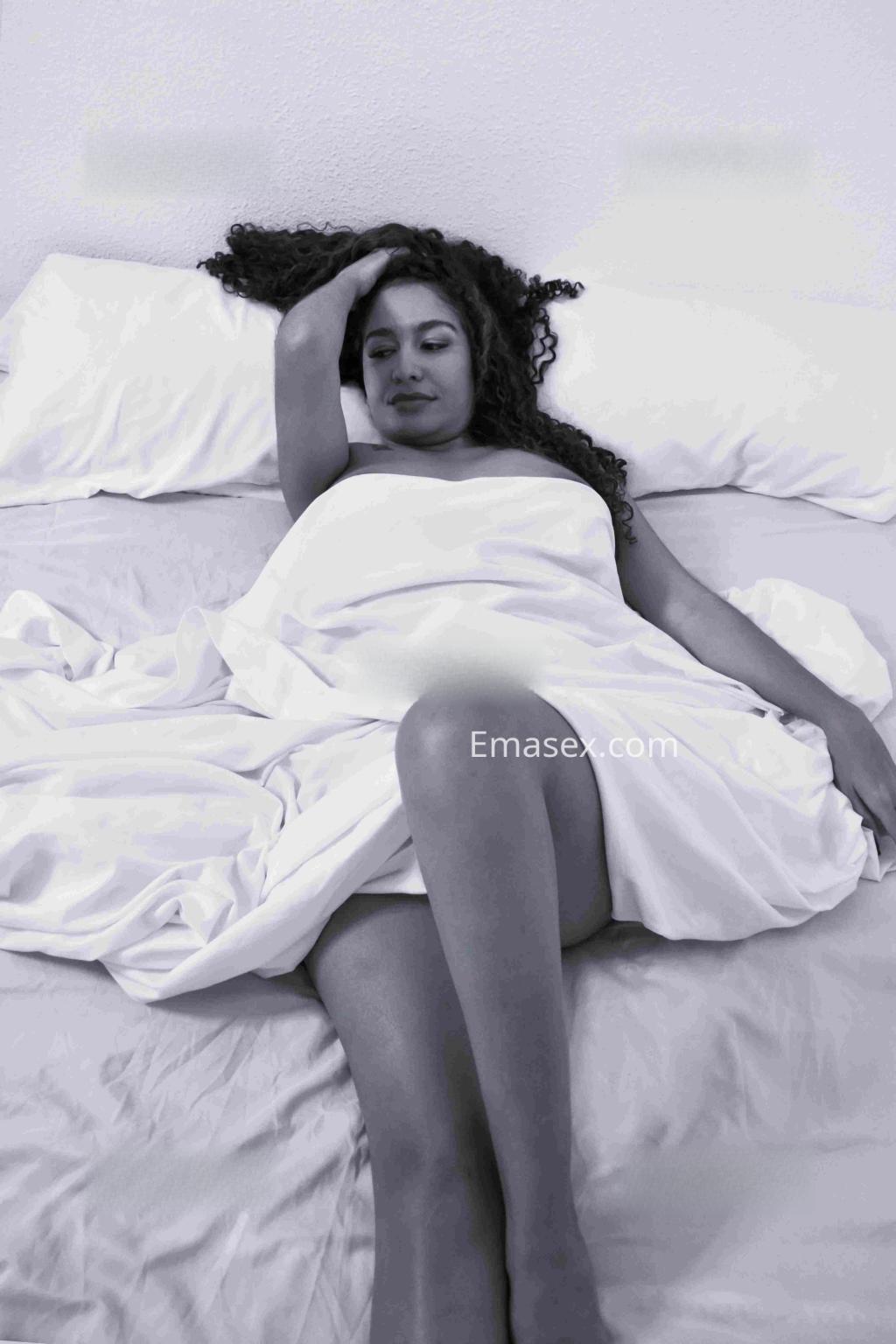 La puta se encuentra recostada en la cama con sábanas blancas, mostrando una pose relajada y sensual en Alicante.
