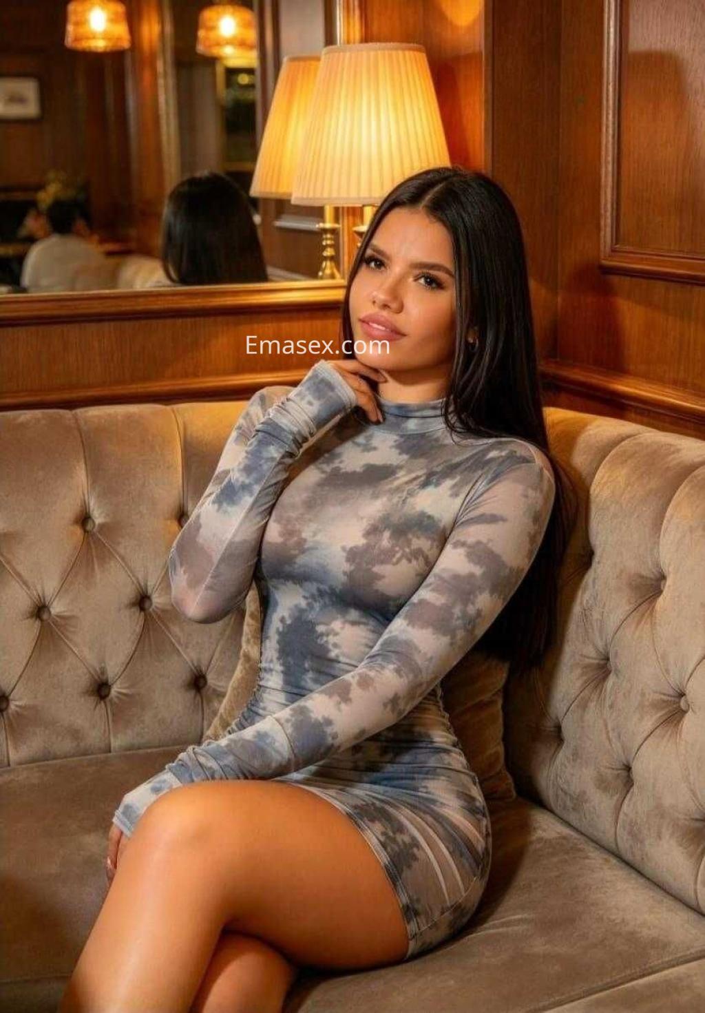 Puta sentada en un sofá de un elegante local en Málaga, vistiendo un vestido ajustado de tonos gris y azul.