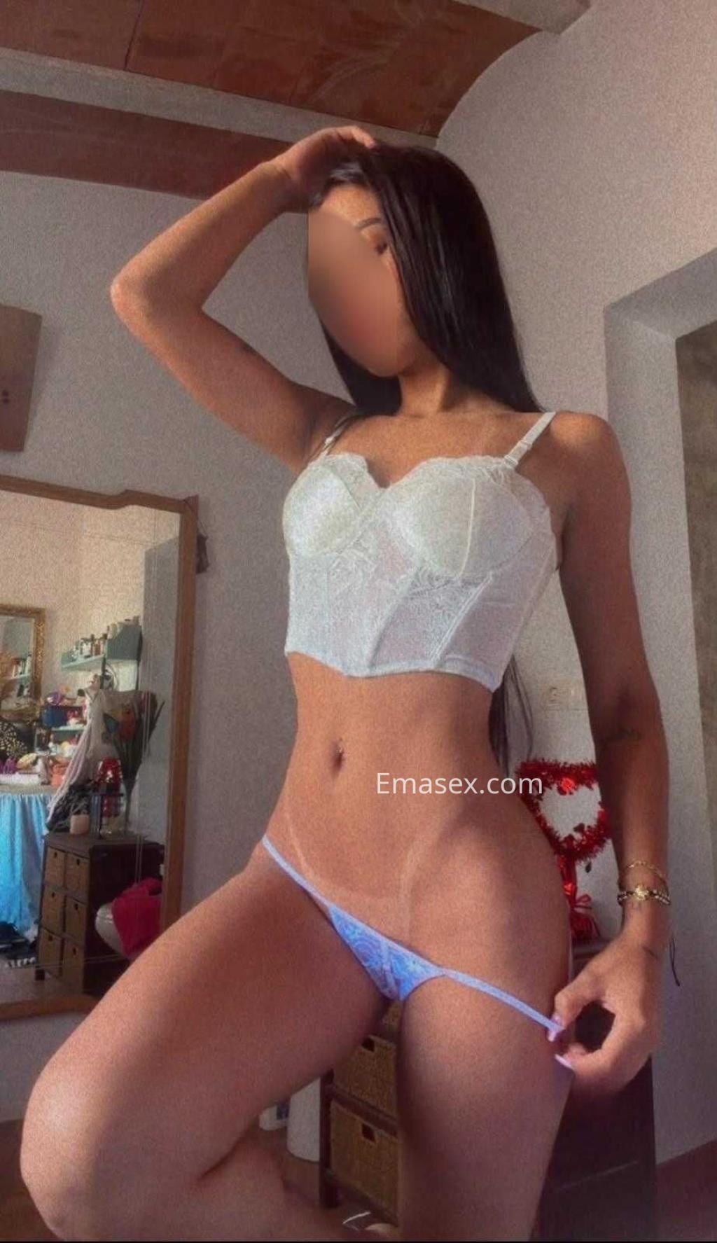 La puta en Alicante luce una blusa blanca de encaje y una tanga azul, posando con confianza en un ambiente interior.