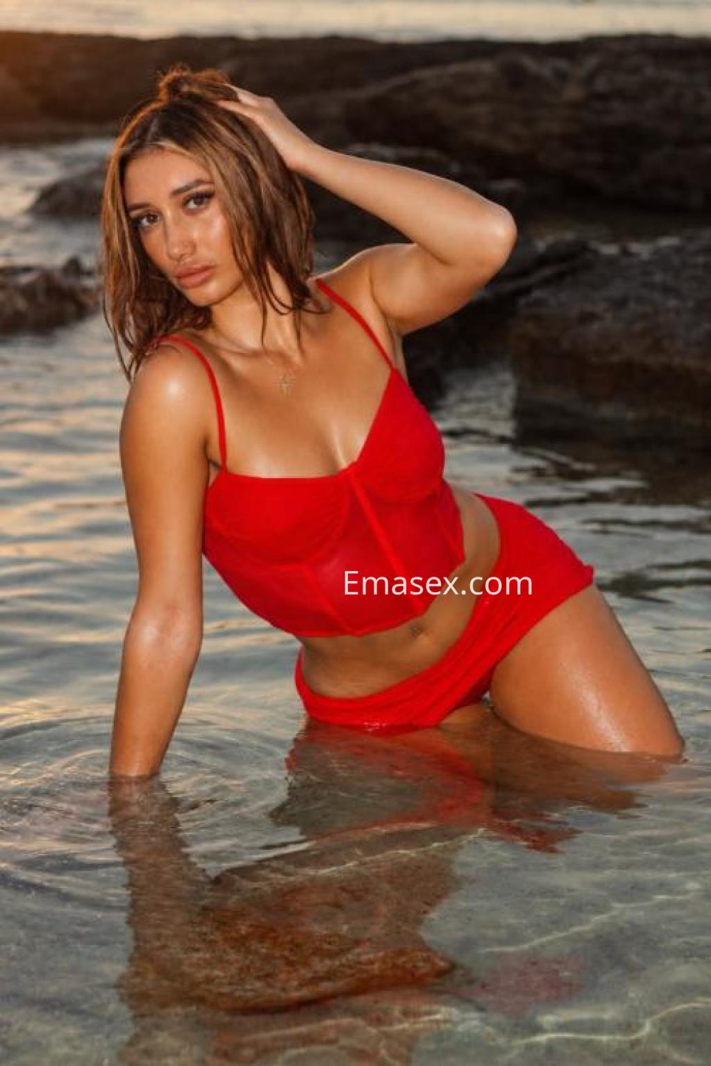 Escort en Ibiza posando en el agua, luciendo un conjunto de dos piezas rojo y cabello suelto, con una expresión relajada.