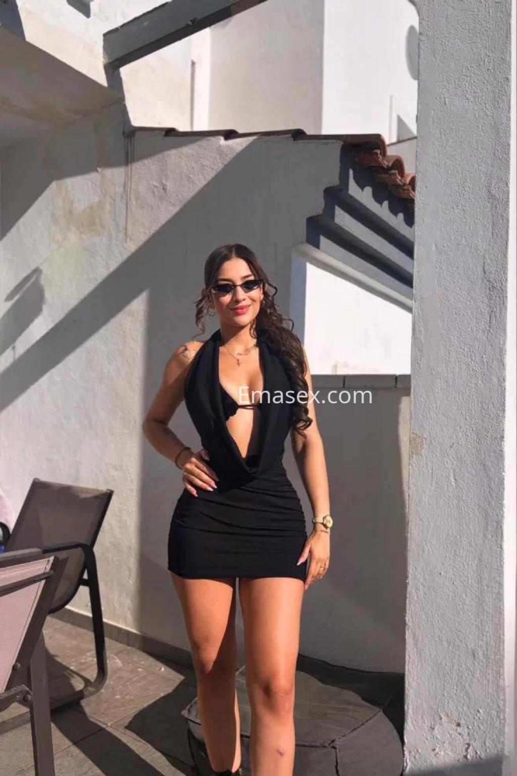 Escort en Cádiz luciendo un vestido negro ajustado y gafas de sol, posando con confianza en un ambiente exterior.