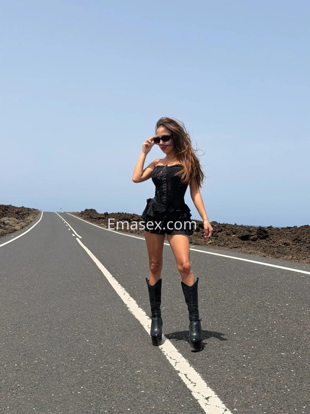 Puta posando en una carretera desierta de Toledo, luciendo un corset negro y botas altas, con gafas de sol.