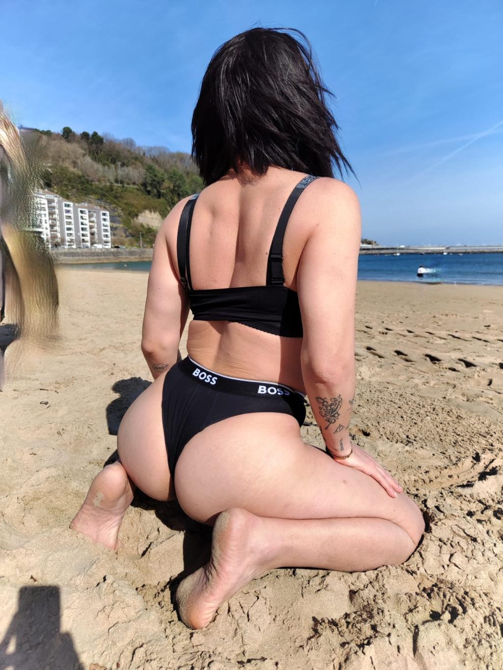 Puta trans de Irún, en la playa, viste un bikini negro y se encuentra en posición de rodillas sobre la arena.
