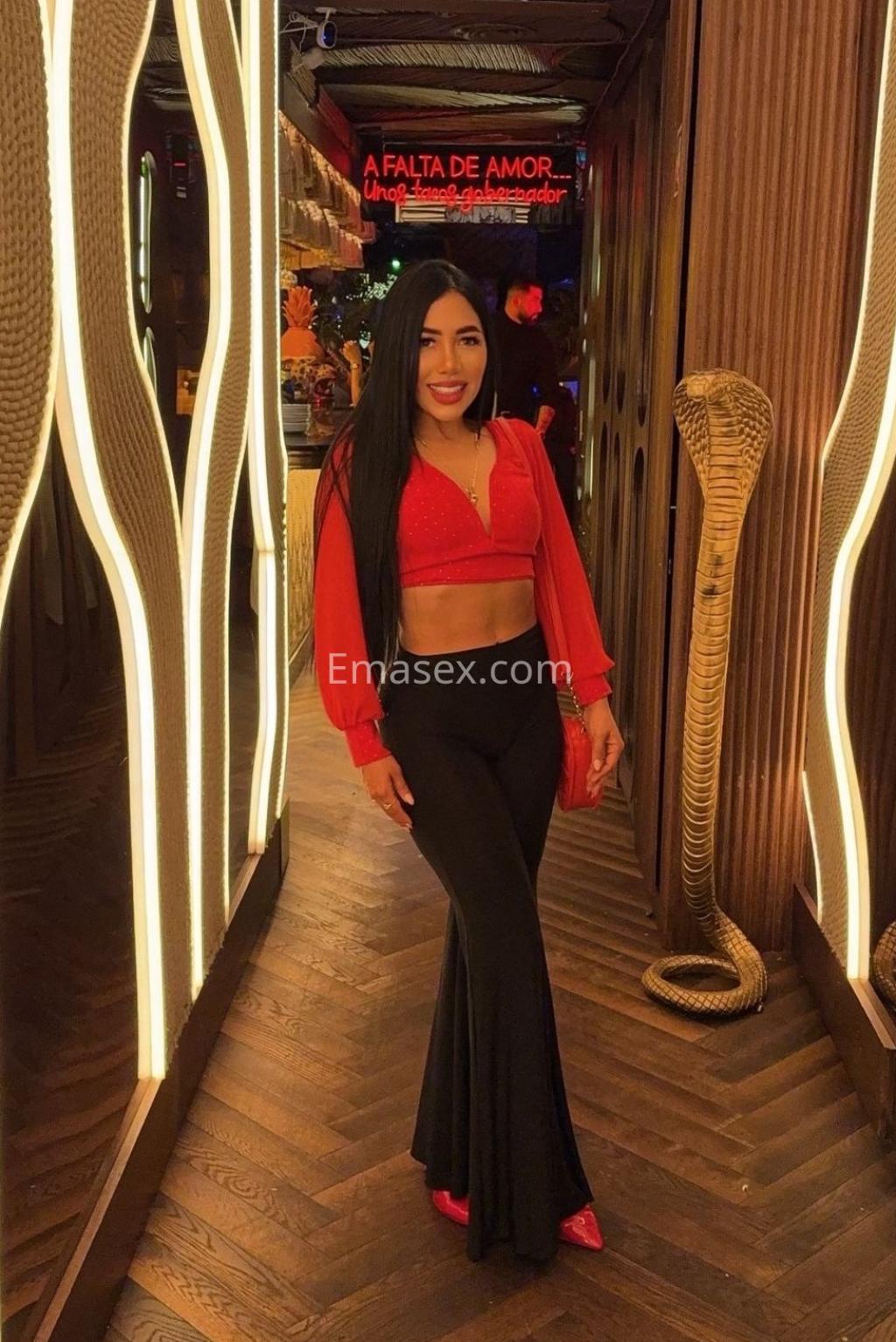 Puta en Albacete con cabello largo y lacio, vistiendo un top rojo y pantalones negros, sonriendo en un ambiente moderno.