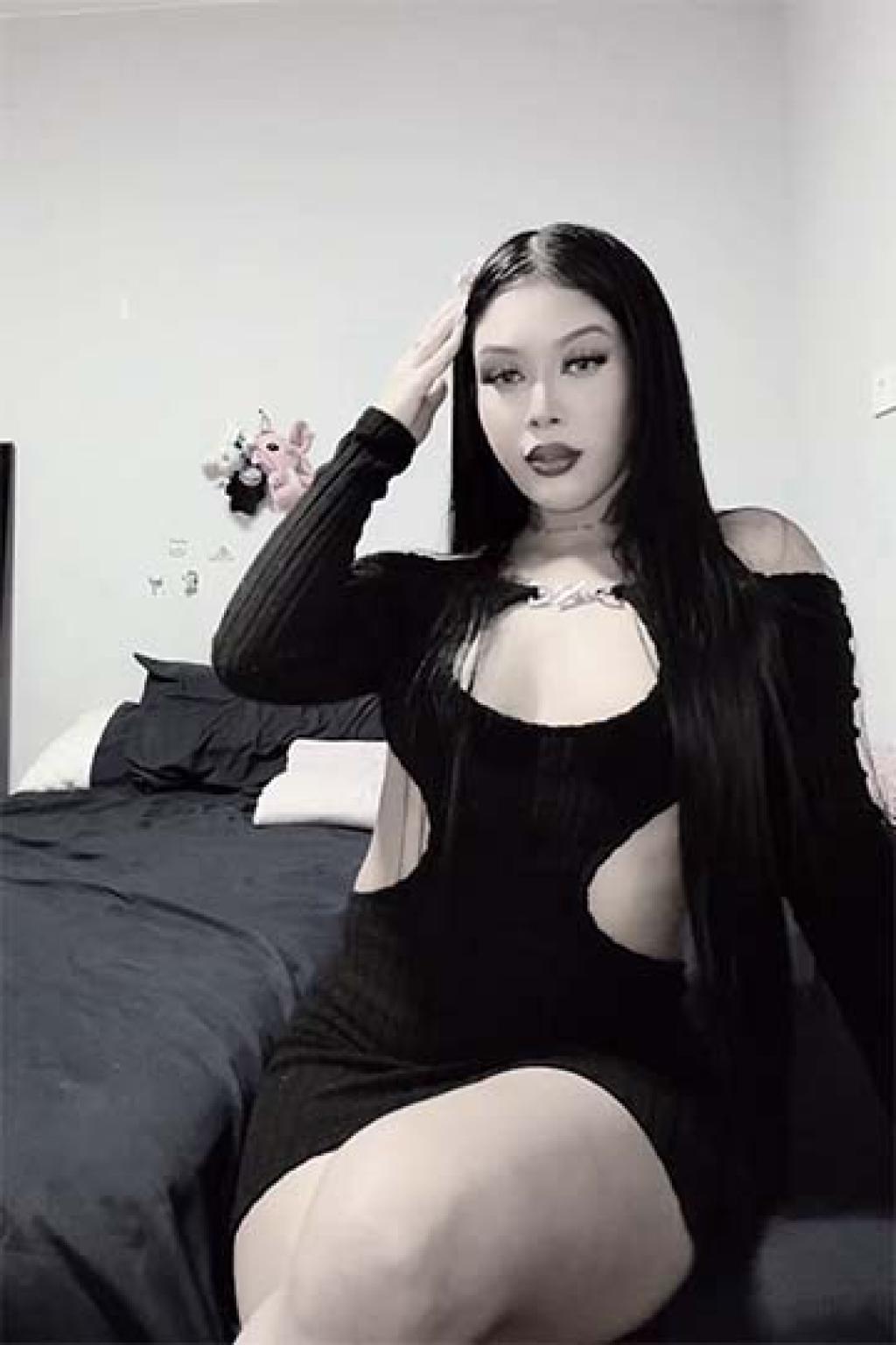 Escort en Logroño con cabello largo y lacio, vistiendo un vestido negro ajustado con cortes laterales, posando en un entorno acogedor.
