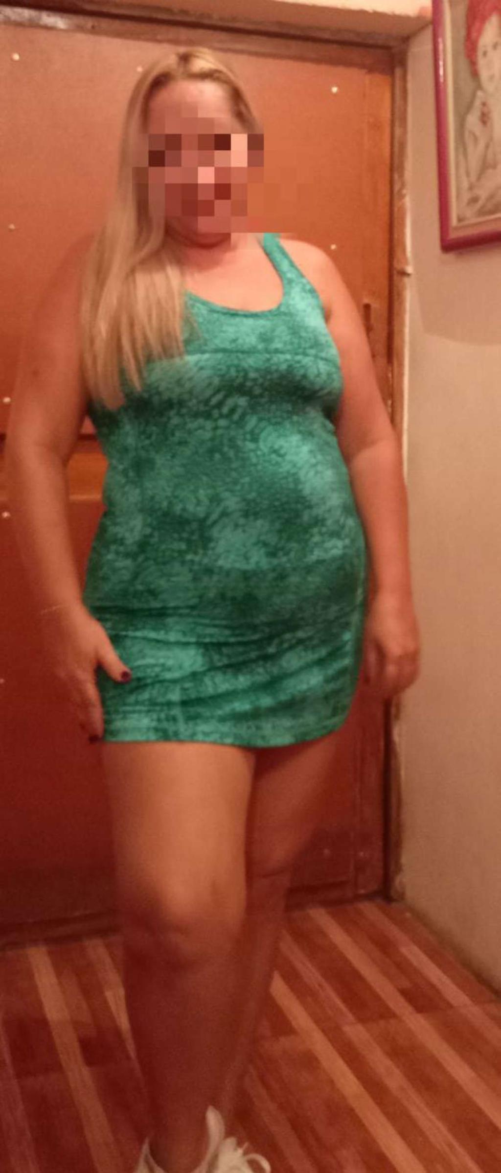 Escort en Vitoria, con vestido verde ajustado y cabello rubio, posando de pie en un entorno interior.
