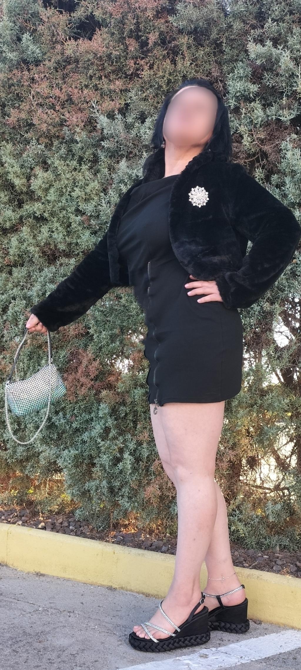 Puta posando en Pamplona con un vestido negro ajustado, chaqueta de pelaje y sandalias de tacón.