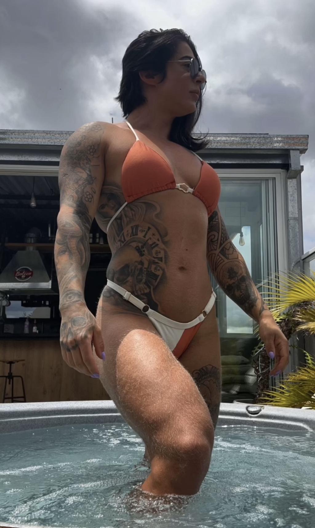 Escort en Bilbao, con cabello oscuro y tatuajes, viste un bikini naranja y blanco mientras posa en una piscina.