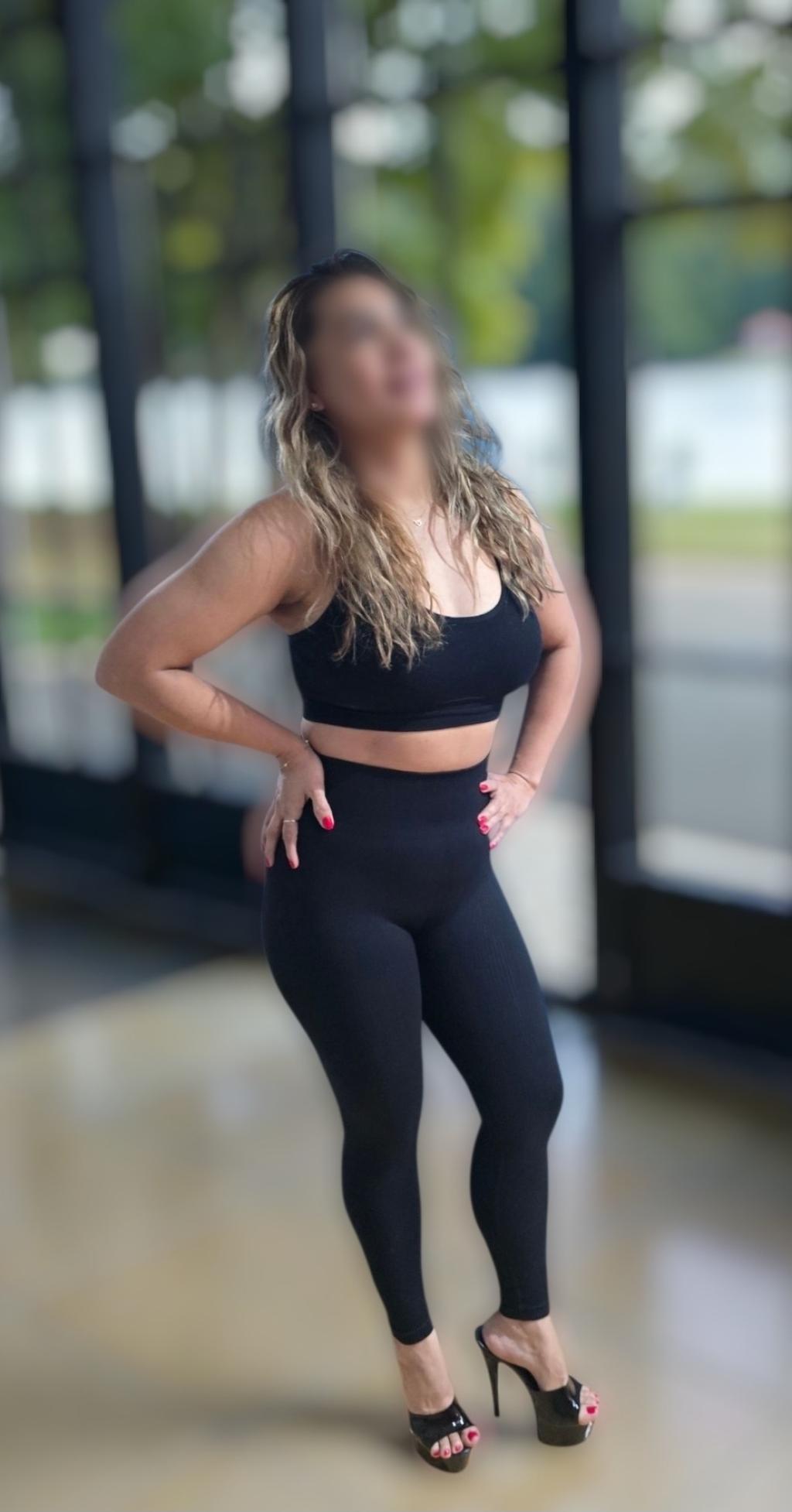 Puta en Vitoria con cabello ondulado, vestida con top negro y leggings ajustados, posando con tacones altos.
