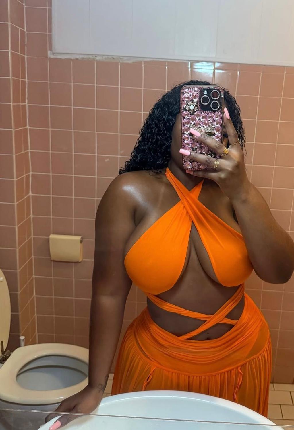 Escort en Pamplona luciendo un top naranja con tirantes cruzados y una falda larga, en un baño con azulejos rosados.