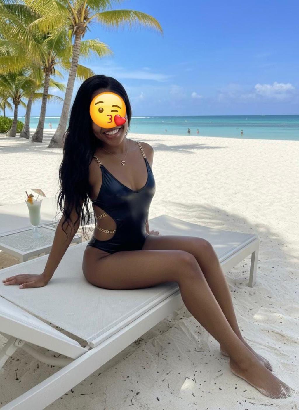 Escort en Ourense con traje de baño negro, sentada en una tumbona frente a la playa y palmeras.
