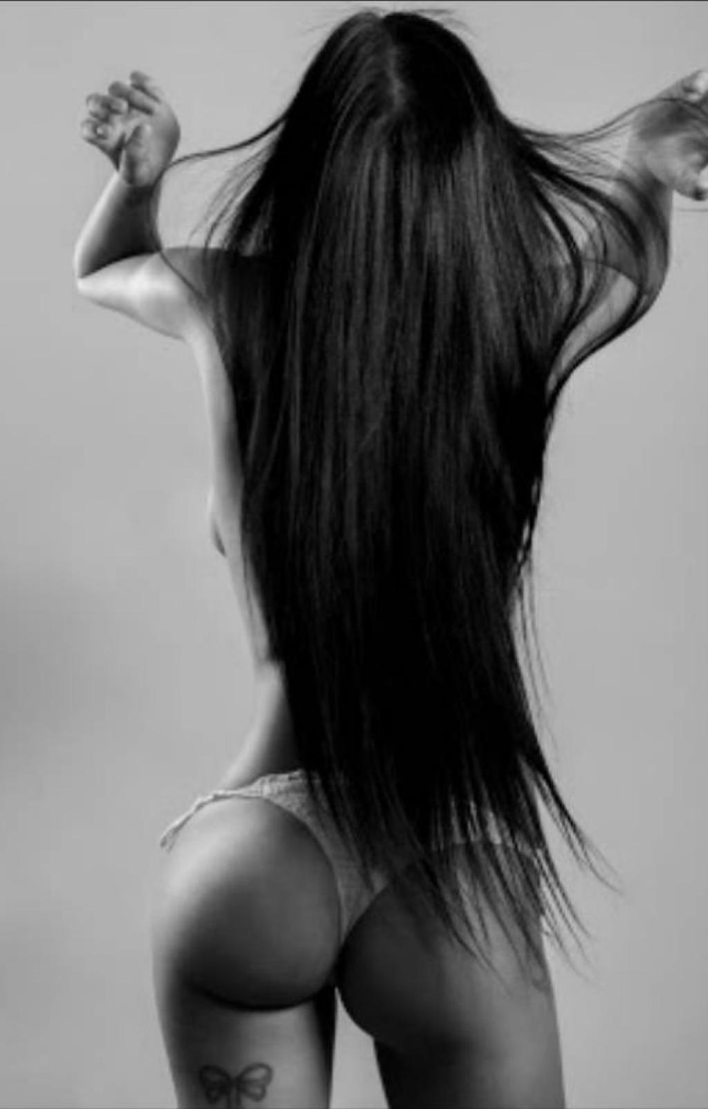 Puta de espaldas con cabello largo y lacio, usando lencería blanca, en un fondo gris, en Sevilla.
