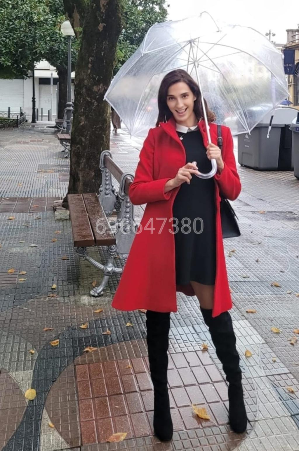 Escort de cabello oscuro y piel clara, viste un abrigo rojo y un vestido negro, sosteniendo un paraguas en Las Palmas.