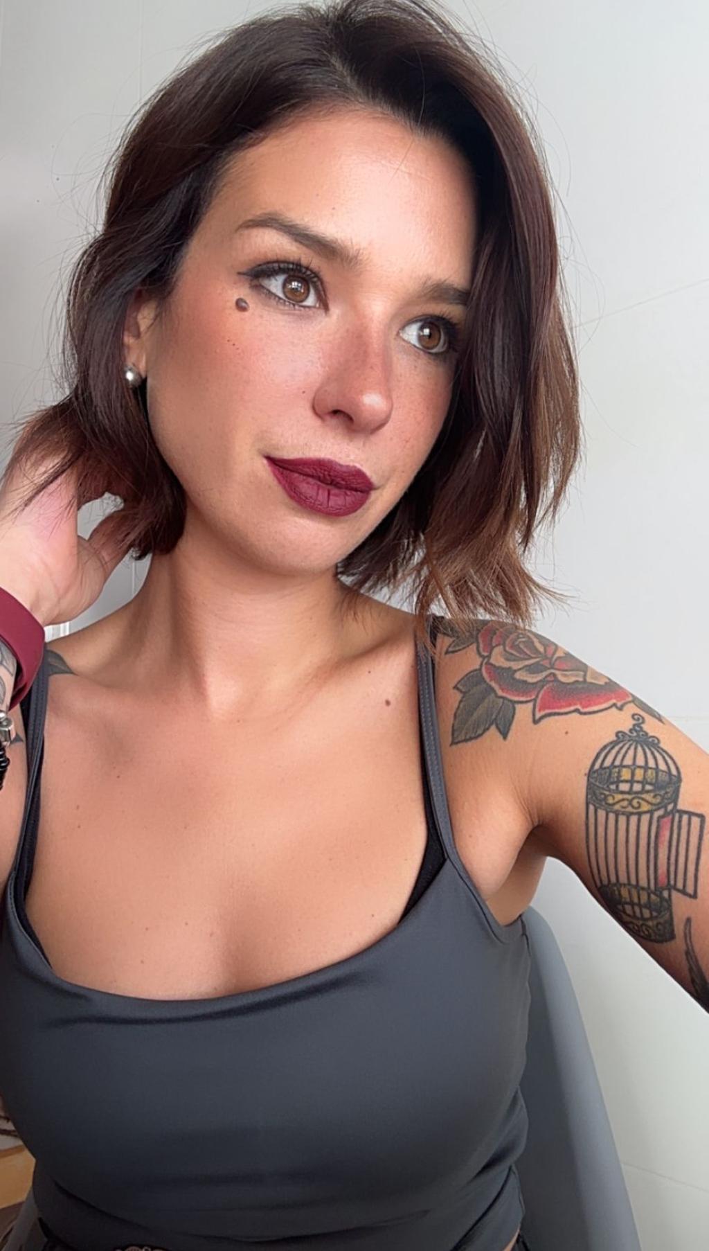 Puta de Gijón con cabello oscuro, blusa sin mangas gris y tatuajes visibles en el brazo, posando sonriente.