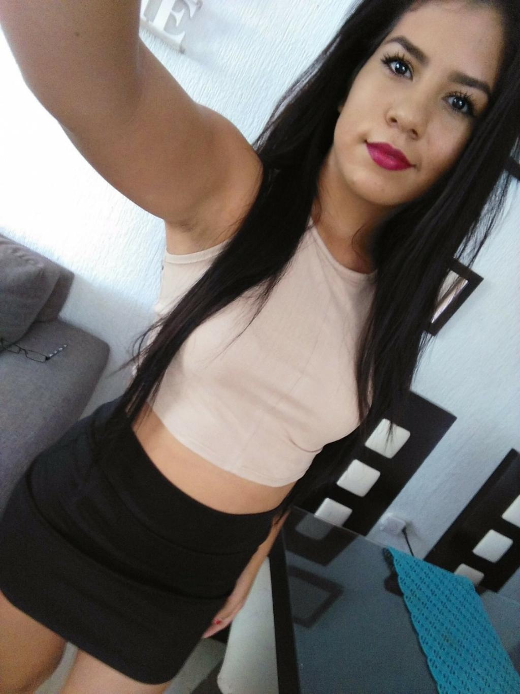 Puta en Burgos con blusa de tirantes beige y falda corta negra, posando con cabello largo y labios rojos.