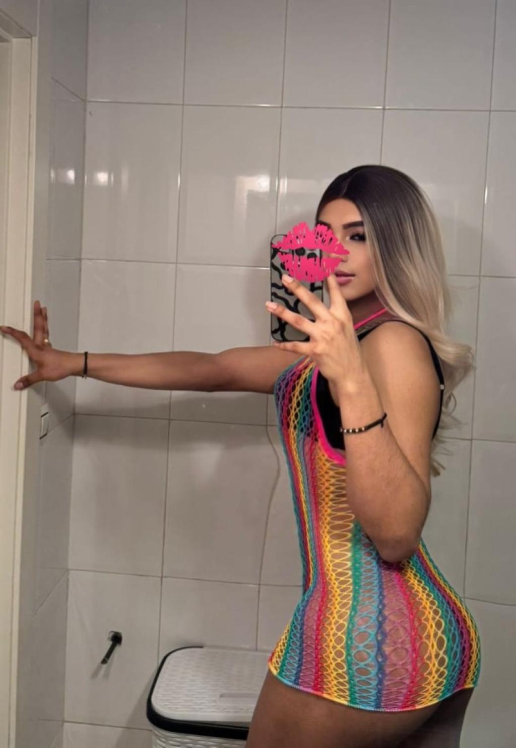 Escort trans en Santander luciendo un vestido de red multicolor en un baño, con cabello largo y rubio.